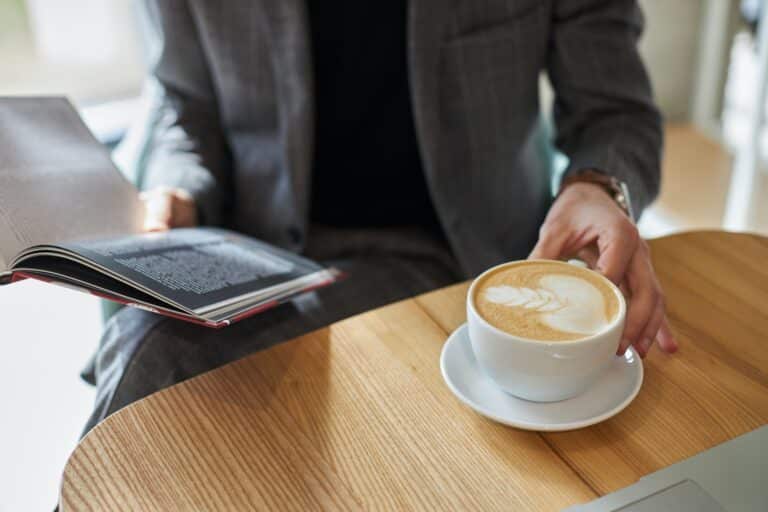 Close-up male designer leafing through a catalogue while sitting at table by a cup of aroma coffee